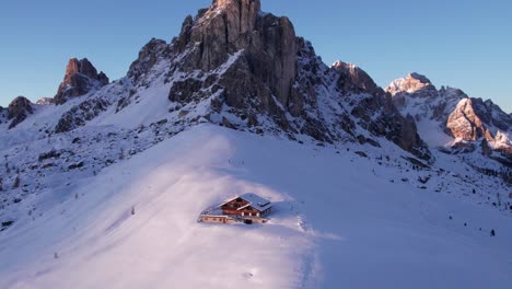 Mountain-lodge-in-winter-snow-in-front-of-Ra-Gusela-mountain-peak,-sunset,-aerial