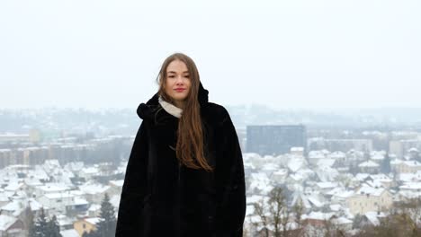 Young-white-female-model-posing-in-front-of-a-city-in-a-black-coat-while-snowing