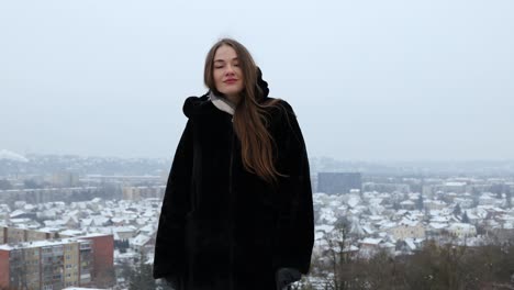 Young-female-model-standing-outside-in-a-black-winter-coat-while-snowing