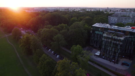 Aerial-flight-showing-golden-sunbeam-lighting-over-park-with-trees-beside-apartment-blocks-in-Krakow-at-sunset