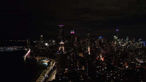In-the-month-of-October,-a-drone-captured-multiple-gloriously-lighted-buildings-or-the-skyline-of-Chicago,-Illinois,-along-with-several-cars-travelling-down-a-highway-and-a-lovely-lake