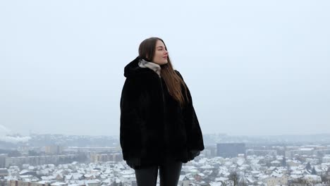 Young-and-attractive-female-model-posing-in-front-of-a-city-in-a-black-coat-while-snowing