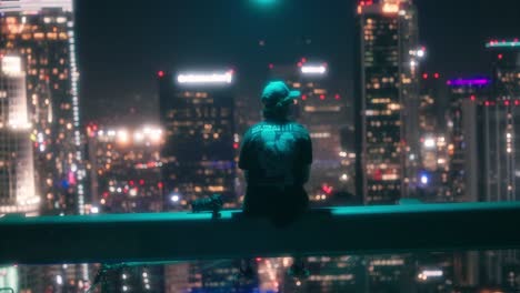 Young-adult-sitting-on-ledge-of-skyscraper-looks-out-at-downtown-Los-Angeles-at-night