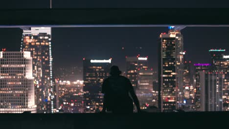 Young-adult-walks-out-on-ledge-of-skyscraper-and-sits-down-to-look-out-at-downtown-Los-Angeles