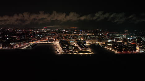 Aerial-view-of-smoke-clouds-moving-over-the-illuminated-Helsinki-cityscape,-winter-evening-in-Finland