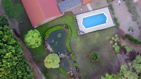 Top-down-drone-shot-of-a-house-with-a-pond,-swimming-pool-and-an-immaculate-stone-garden-with-surrounding-green-hedges