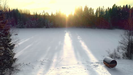 Aerial-drone-backward-moving-shot-of-bright-sunrise-from-behind-coniferous-trees-over-frozen-lake-alongside-barrel-sauna