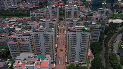 Dramatic-drone-Fly-in-over-central-road-between-tall-buildings-of-a-massive-symmetrical-housing-estate-on-sunny-day-featuring-river,-towers,-rooftops,-gardens,-swimming-pool-and-tennis-courts-1