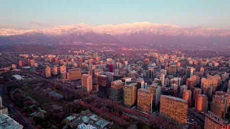 Aerial-parallax-of-Las-Condes-financial-district-buildings,-snow-capped-mountains-in-background-at-sunset,-Santiago,-Chile