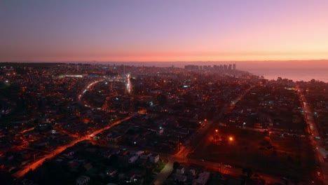 Aerial-parallax-of-Concon-neighborhood-buildings-illuminated-at-night-near-sea-shore-at-colorful-golden-hour,-Valparaiso,-Chile