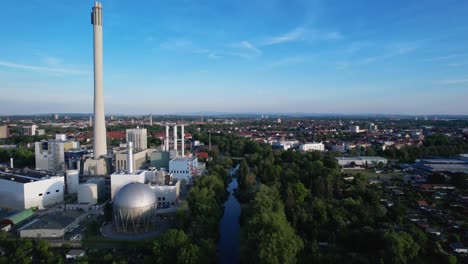Large-round-storage-tank-with-a-high-white-chimney-of-a-large-power-station-in-the-German-city-of-Braunschweig-on-the-river-Oker-with-a-clear-blue-sky-in-the-background