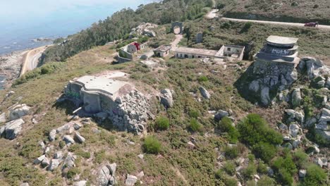 Aerial-drone-rotating-shot-of-old-abandoned-army-bunkers-with-cannon-along-hilly-terrain-overlooking-the-beautiful-blue-sea-at-daytime