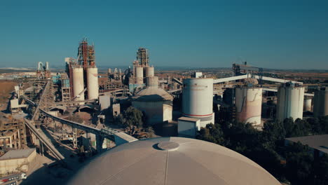 Aerial-shot-of-a-huge-cement-factory,-innovative-industrial-area-with-lots-of-hangars-and-trucks,-beautiful-area-with-trees,-parallax-shot-1