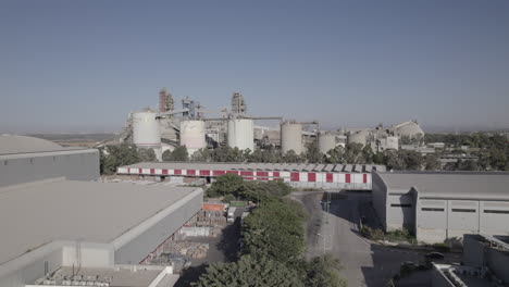 Aerial-shot-of-a-huge-cement-factory,-an-innovative-industrial-area-with-lots-of-hangars-and-trucks,-a-beautiful-area-with-trees,-push-in-shot