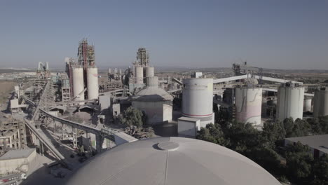 Aerial-shot-of-a-huge-cement-factory,-innovative-industrial-area-with-lots-of-hangars-and-trucks,-beautiful-area-with-trees,-parallax-shot