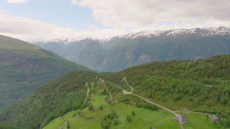 Casas-Y-Carretera-Escénica-En-Montaña-Verde-Con-Cadenas-Montañosas-Cubiertas-De-Nieve-En-La-Distancia-En-Noruega