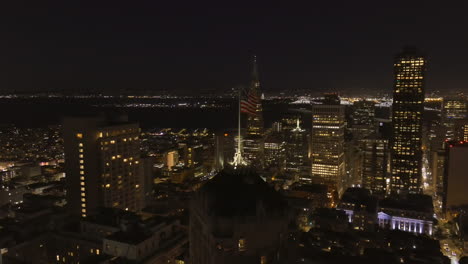 A-United-States-flag-blowing-at-nighttime-on-a-tall-building-with-the-San-Francisco-city-skyline-as-a-backdrop---aerial-parallax-edited-at-the-end-for-a-transition