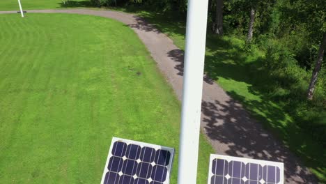 Park-pole-with-solar-panels-reflecting-sunlight-and-windmill-on-top,-close-up-aerial-ascend
