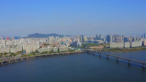 Drohne-Schoss-An-Einem-Sonnigen-Tag-über-Den-Fluss-Han-In-Richtung-Eines-Wohngebiets-Und-Einer-Brücke-In-Der-Stadt-Seoul