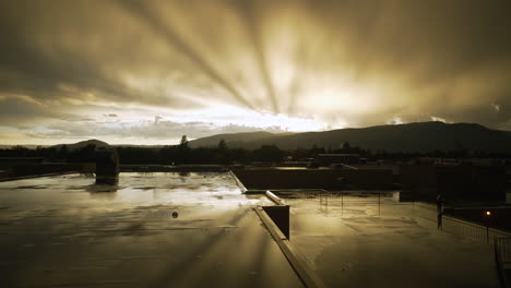 Las-Gotas-De-Lluvia-Caen-Sobre-Los-Charcos-Formados-En-Un-Techo-Mientras-El-Sol-Se-Pone-Bellamente-Detrás-De-Las-Montañas-Distantes,-Creando-Rayos-Divinos-Desde-Las-Nubes