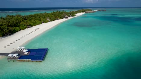 Aerial-view-of-maldives-island-beach-with-overwater-pool-lounge-chairs-palm-trees-bungalows-by-shallow-clear-blue-water