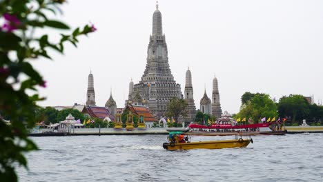 The-charming-river-of-Bangkok-with-longtail-boats-sailing-on-the-river,-with-the-impressive-Wat-Arun-in-the-background,-during-daytime-in-Thailand
