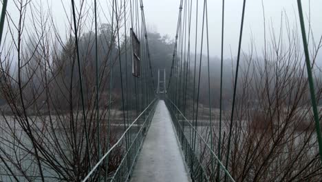 Drone-ascending-to-the-top-of-hanging-walkway-over-the-river-with-misty-forest-in-the-distance---Drone-view