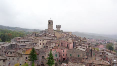 Medieval-town-on-hill-in-Tuscany-countryside-with-tall-church-bell-tower,-cloudy-day,-aerial