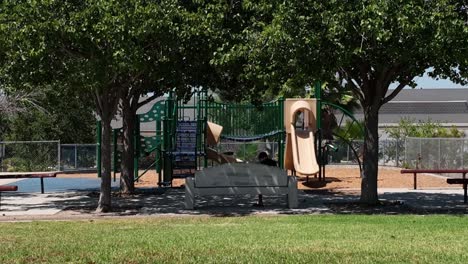 Young-Guy-Person-Sitting-Alone-on-Bench-Under-Trees-in-Schoolyard-Playground-Park-next-to-Green-Lawn-Field,-Lonely-Recreation-and-Relaxing-in-Nature-in-Summer,-Backward-View