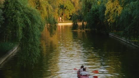 Slow-motion-fixed-shot-of-a-couple-in-a-red-kayak-on-an-urban-lake-at-sunset-with-trees-on-the-banks