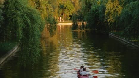 Toma-Fija-En-Cámara-Lenta-De-Una-Pareja-En-Un-Kayak-Rojo-En-Un-Lago-Urbano-Al-Atardecer-Con-árboles-En-Las-Orillas