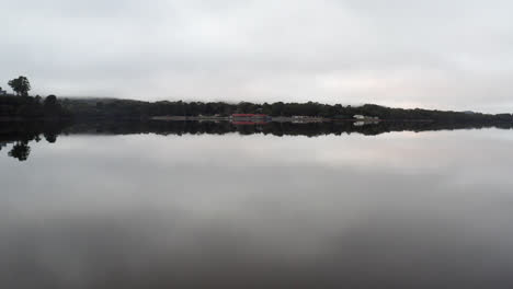 Antena:-Drone-Volando-A-Baja-Altura-Sobre-Un-Lago-Perfectamente-Quieto-Hacia-La-Costa-Cerca-De-Strahan,-Tasmania