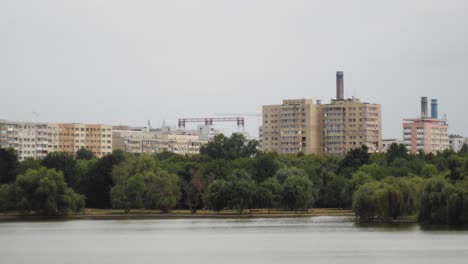 A-pan-right-slow-motion-shot-reveals-an-urban-landscape-of-Titan-Park-in-Bucharest-with-a-lake,-some-trees,-and-communist-buildings
