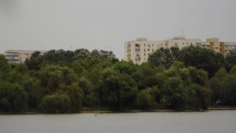 A-pan-right-tight-slow-motion-shot-reveals-an-urban-landscape-of-Titan-Park-in-Bucharest-with-a-lake,-some-trees,-and-communist-buildings