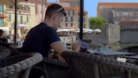 Young-male-entrepreneur-working-online-at-a-restaurant-while-drinking-a-coffee-and-virgin-cocktail-in-Collioure,-France