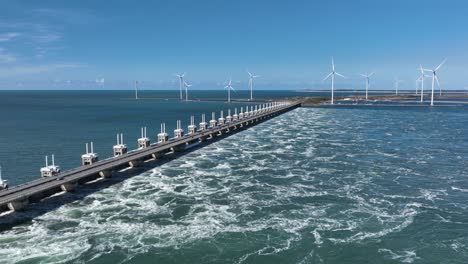 Slow-aerial-forward-shot-of-Oosterschelde-storm-surge-barrier-at-Delta-Works-and-rotating-wind-turbines-in-background-during-sunny-day-with-blue-sky---Netherlands,Europe