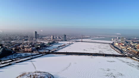 Drone-backwards-shot-over-iced-and-snowy-Daugava-River-in-Riga-City-with-bridges-and-buildings