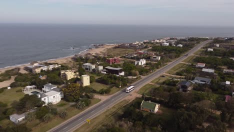 Aerial-tracking-shot-of-white-bus-driving-by-coastal-road-in-Maldonado-Town,Uruguay---Beautiful-landscape-with-beach-and-ocean-water-during-sunlight