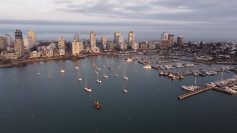 Aerial-view-of-port-with-anchored-sailing-boats-in-front-of-Punta-del-Este-City-at-sunset---backwards-flight