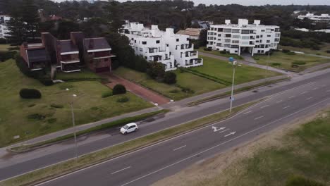 Aerial-tracking-shot-of-white-car-driving-on-boulevard-road-in-Punta-del-Este,-Uruguay---Luxury-modern-apartment-buildings-beside-beach