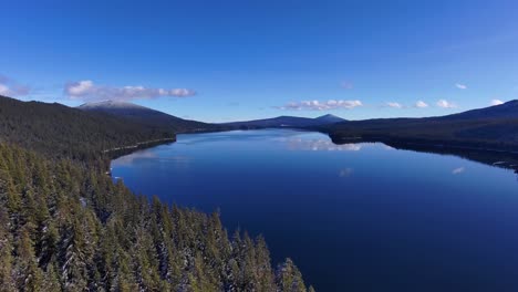 Wide-drone-shot-above-snowy-trees-and-clear-lake-on-sunny-winter-day,-60fps