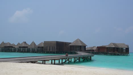 Edificios-Sobre-El-Agua-En-El-Complejo-De-Maldivas-Con-Agua-Azul-Y-Cielo-Despejado