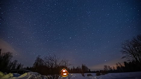 Static-shot-of-a-wooden-cabin-surrounded-by-thick-layers-of-snow-with-stars-moving-through-sky-in-timelapse-at-night-time