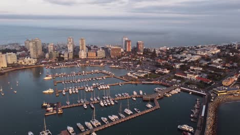 Aerial-backwards-flight-showing-beautiful-skyline-of-Punta-del-Este-City-with-Port-Harbor-and-parking-sailing-boats-during-golden-sunset---South-Atlantic-Ocean-in-background-overcast-with-dark-clouds
