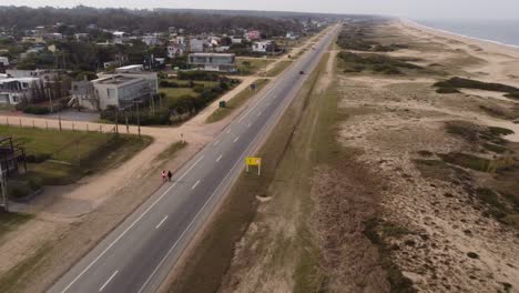 Aerial-tracking-shot-of-two-women-walking-at-side-of-coastal-road-near-beach-in-Maldonado,Uruguay