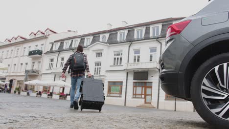 Man-with-luggage-closing-trunk-of-car-and-walking-away-towards-white-tenement