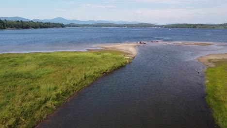 Aerial-drone-footage-over-a-channel-leading-into-Lovewell-Pond-in-Maine,-with-kayakers-on-a-sandbar-and-mountains-in-the-distance