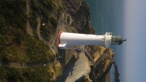 A-reversing-vertical-drone-shot-of-a-person-jogging-along-a-wooden-pathway-away-from-a-beautiful-white-lighthouse