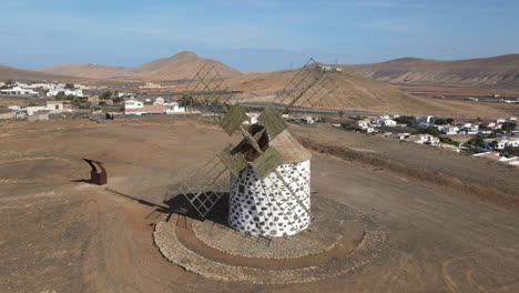 Aerial-shot-in-orbit-over-an-old-cereal-mill-on-the-island-of-Fuerteventura,-displaying-the-blades-of-the-mill-and-the-mountains-around-Villaverde