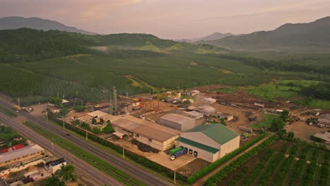 Aerial-flyover-traffic-on-highway,-industrial-factory-and-green-Coconut-Plantation-at-dawn-in-the-morning---VIlla-Altagracia,DOminican-Republic